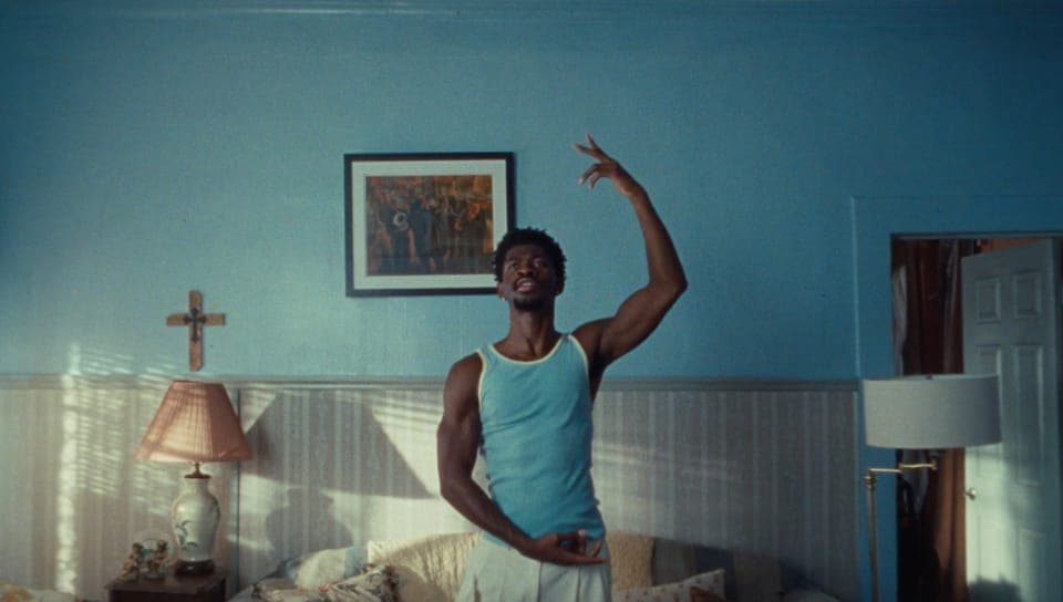 The image shows a man standing in a room with blue walls. He is wearing a blue tank top and white shorts, and is making a peace sign with his right hand. The room has a vintage aesthetic, with a bed featuring a white comforter and pillows, a lamp with a cream-colored shade, and a framed picture on the wall. A white doo