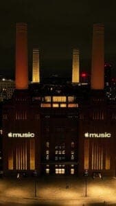 The image depicts the iconic Apple Music building, a renowned landmark in London, UK. The building is illuminated by warm, yellowish lights, creating a striking contrast against the dark night sky. The building's facade features a series of tall, cylindrical structures, reminiscent of smokestacks, and is adorned with t
