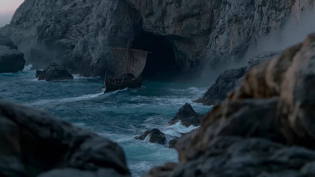 The image depicts a dark-colored, wooden Viking longship sailing towards a cave entrance in a rocky coastline. The ship is moving from left to right, leaving a trail of white foam in its wake. The cave entrance is a dark opening in the rocky cliff face, and the surrounding area is filled with rocks and boulders. The wa