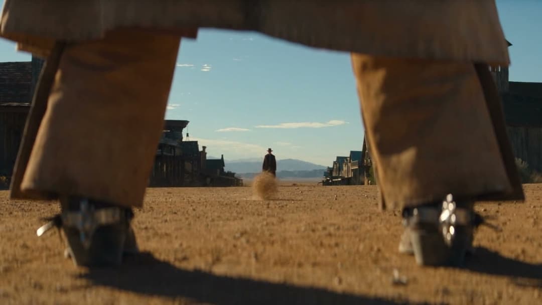 The image shows a person wearing tan or beige pants and boots, standing on a dirt road in a desert-like setting. The person is facing away from the camera, with their feet slightly apart. In the distance, a lone cowboy is riding a horse, kicking up a cloud of dust. The background features a row of buildings, possibly a