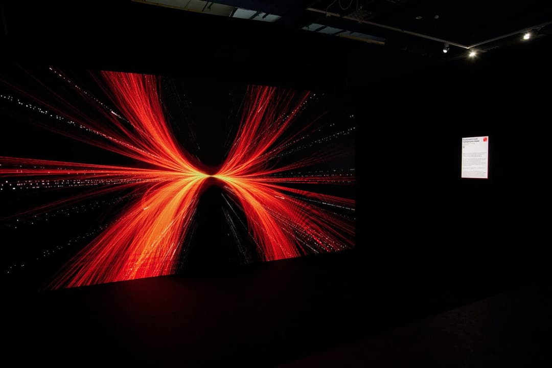 The image depicts a dimly lit room with a large, rectangular screen displaying a vibrant, abstract image of a starburst or cosmic event. The image is composed of bright orange and red lines radiating outwards from a central point, creating a sense of movement and energy. The screen is mounted on a black wall, and a sma