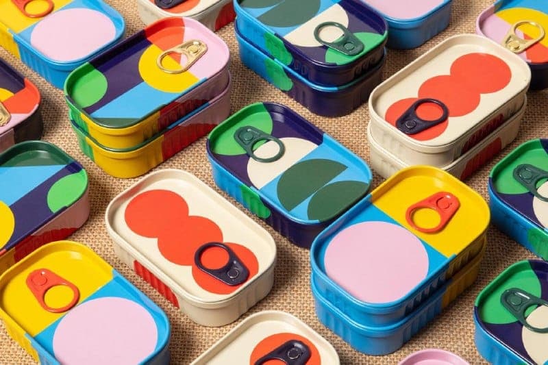 The image shows a collection of colorful cans arranged on a beige background. The cans are stacked in various configurations, creating a visually appealing pattern. Each can features a unique design, with a combination of geometric shapes and solid colors. The colors include blue, yellow, pink, green, and orange, with