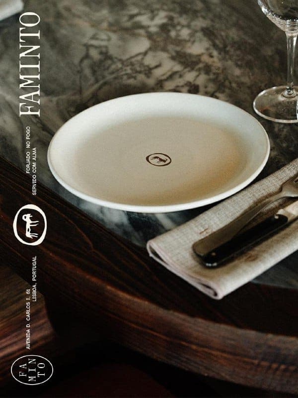 The image shows a table setting with a white plate featuring a brown logo in the center. The plate is placed on a dark brown wooden table. To the right of the plate, there is a folded beige napkin and a knife with a black handle. A wine glass is visible in the background. The table is set against a marble-like surface
