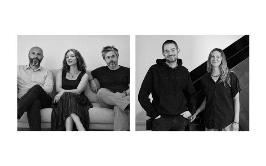 This black and white photograph captures four individuals, two men and two women, in a casual setting. The two men are seated on a couch, one wearing a light-colored shirt and the other a dark-colored hoodie. The two women are standing, one wearing a dark-colored top and the other a dark-colored top with a patterned sk