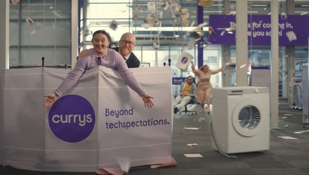 The image shows a lively scene in a store, possibly a Curry store, with a large white tent-like structure in the foreground. The tent features a purple logo with the word "currys" written in white. Two individuals are standing behind the tent, one wearing a dark suit and the other a light purple/lavender shirt. The ten
