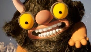The image shows a close-up view of a whimsical, cartoonish monster with a large, round, yellow eye and a pink nose. The monster has a dark brown or black fur-like texture and a wide, open mouth revealing a set of white teeth. Its left hand is extended, palm up, as if reaching out. The monster is wearing a reddish-orang