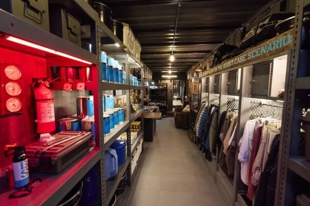 The image depicts a narrow, well-lit corridor with a high ceiling and exposed metal beams. The walls are lined with shelves that hold a variety of items, including blue cans, red fire extinguishers, and other miscellaneous objects. The floor is covered in a gray carpet. On the right side of the corridor, a row of metal