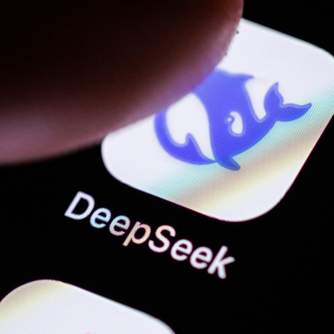 The image shows a close-up view of a smartphone screen displaying the app icon for "DeepSeek". The icon is a blue dolphin swimming against a white background. The word "DeepSeek" is written in white text below the icon. The phone's screen is black, and the image is taken from a slightly elevated angle, with the top lef