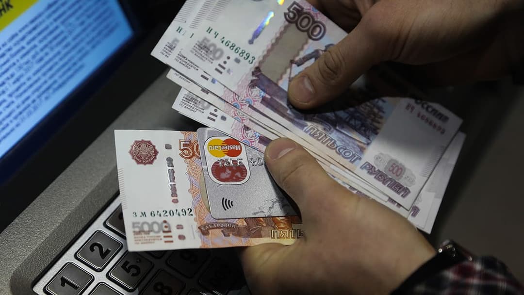 The image shows a person's hands holding a stack of Russian 500 rubles banknotes and a credit card. The banknotes are predominantly white with pink and red accents, and the numbers "500" and "������������" are visible. The credit card is a silver/grey color with a red and white logo. The person is wearing a dark-colore