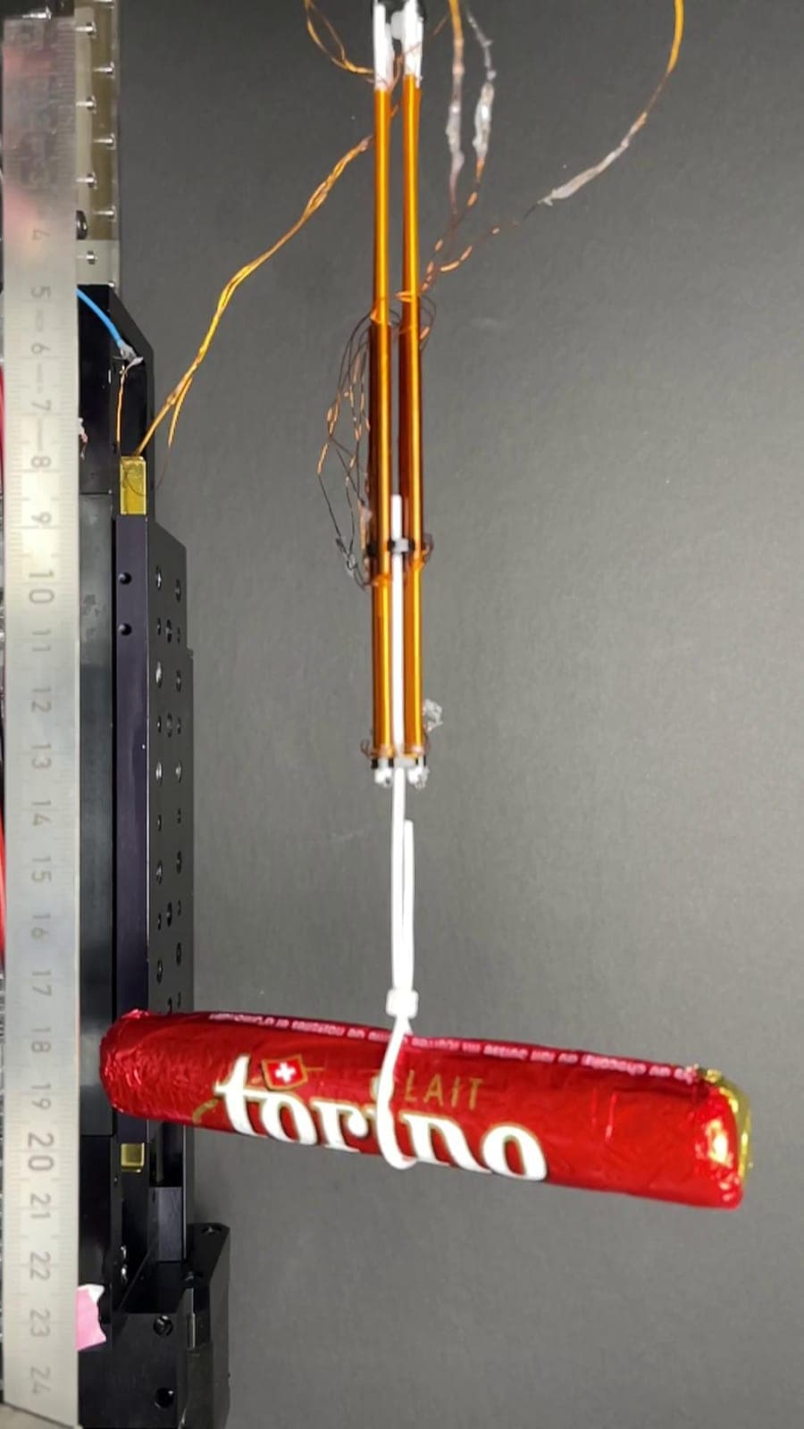The image shows a black rectangular device with a series of small holes or openings, possibly for measurement or calibration. A red and white Torino cheese stick is suspended from a white wire, which is attached to a black wire that extends upwards and outwards. The wires are connected to a black rectangular device wit