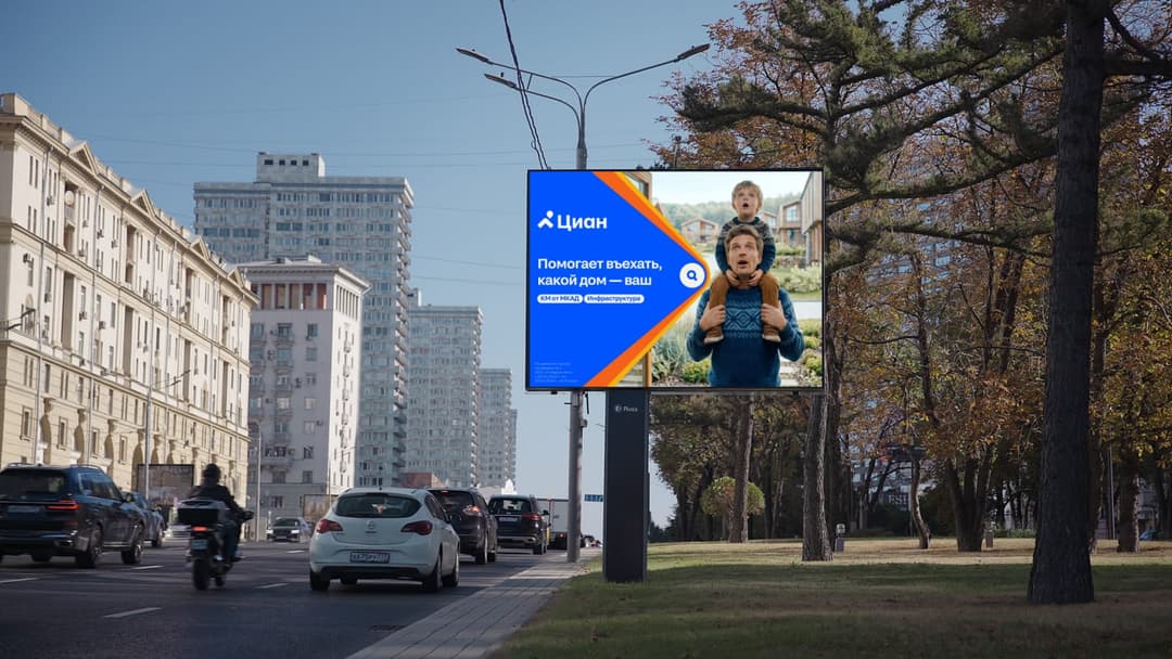 The image shows a large billboard in a city setting. The billboard is primarily blue and orange, with a white background. It features a cartoon image of a man carrying a child on his shoulders. The billboard is situated on a pole on the side of a street, with a sidewalk and a grassy area nearby. The street is lined wit