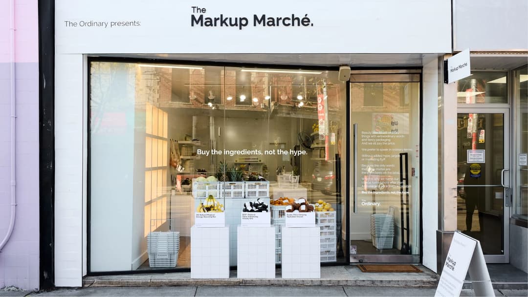 The image shows the storefront of "The Markup Marche", a store that sells ingredients. The store's name is prominently displayed in white letters on a black sign above the entrance. The entrance is marked by a glass door, and a sign above it reads "The Ordinary presents". The store's interior is visible through the gla
