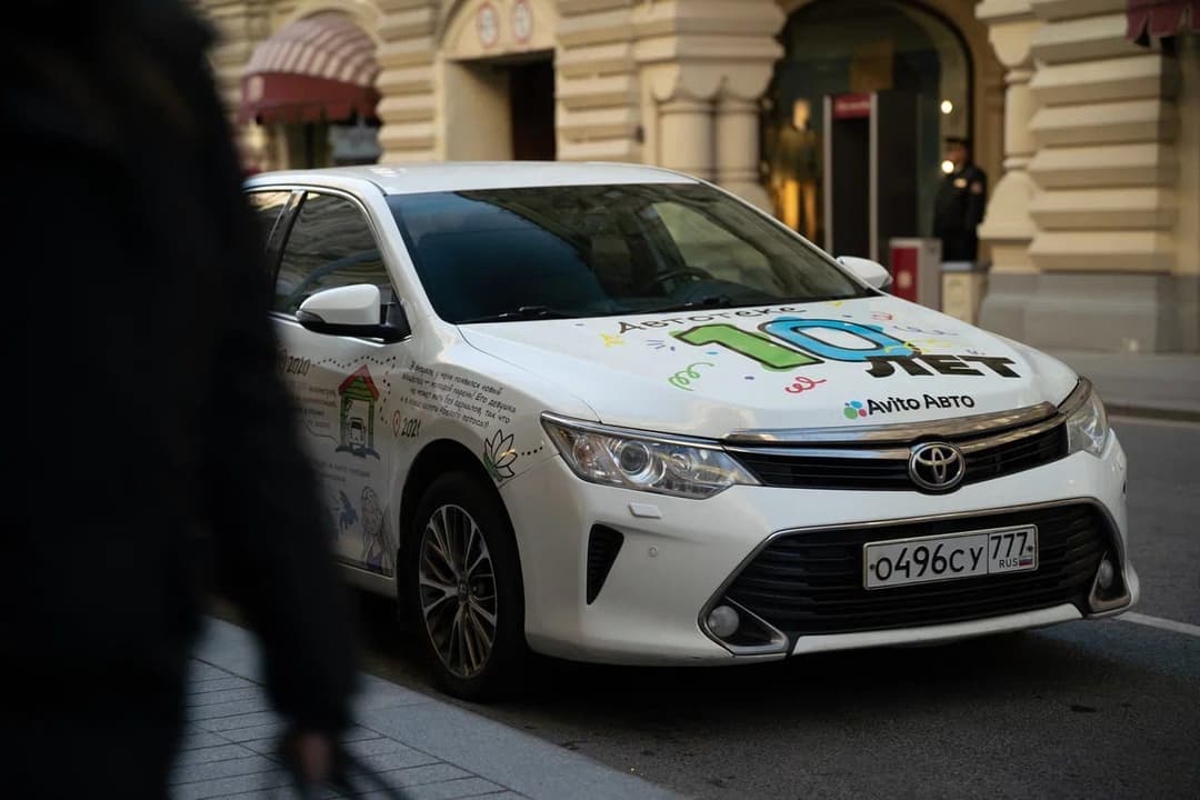 A white Toyota Camry parked on a city street, with a hand-drawn "10 лет" (10 years) and "Avito Авто" logo on its hood and a Russian license plate. Its sides are covered in doodles, and a building with arched windows and columns is visible behind it.