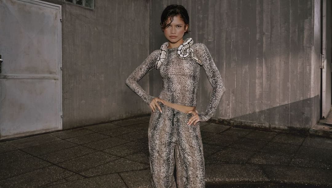 The image shows a young woman standing in front of a gray concrete wall. She is wearing a snakeskin-patterned outfit consisting of a top and pants, with a snake design on the top and a snake-like pattern on the pants. The woman has dark brown hair and is looking directly at the camera. Her hands are on her hips, and sh