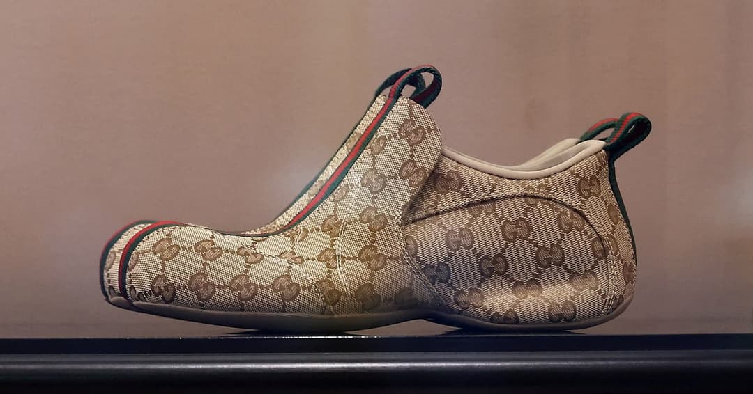 The image shows a pair of Gucci shoes displayed on a black shelf against a beige wall. The shoes are primarily beige or cream-colored with a brown Gucci pattern and a red and green striped trim. The shoes have a pointed toe and a low heel, and they feature a strap across the top of the foot. The shoes are positioned in
