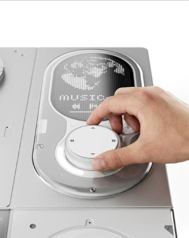 The image shows a hand interacting with a white and silver device, possibly a music player or similar audio device. The device has a circular control panel with a white knob and a digital display that shows a world map. The word "Music" is also visible on the display. The device is set against a white background, and t