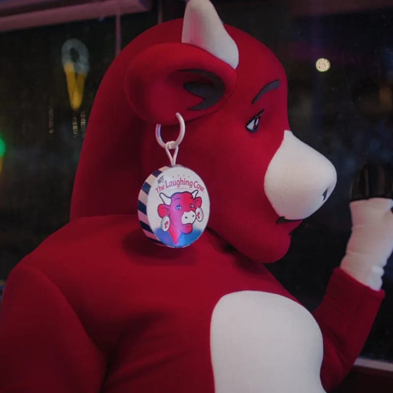 The image shows a red and white plush toy cow, possibly a mascot, wearing a white and red striped shirt. The cow is positioned in profile, facing towards the right side of the image. It has a white nose and a pink face, and its ears are large and pointed. The cow is wearing a white and red striped earring with a white