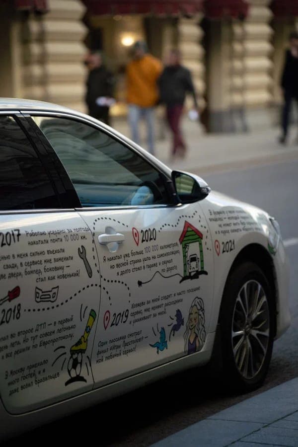 Side view of a white Toyota Camry covered in hand-drawn doodles and timeline text with years 2017 to 2021, dotted route lines, and red location pins. The car is on a city street with blurred pedestrians in the background.