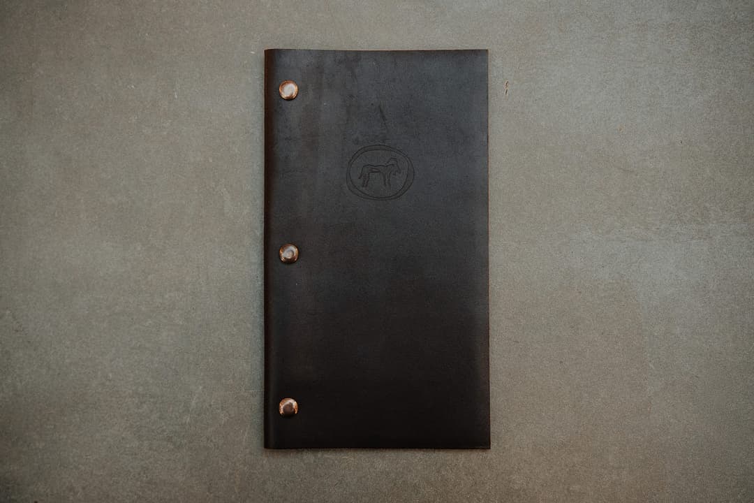 The image shows a black leather menu holder resting on a gray concrete surface. The menu holder has three silver-colored metal rivets on the left side, and a circular logo featuring a stylized animal, possibly a dog or wolf, is centered on the front. The menu holder is positioned in the center of the image, with the gr