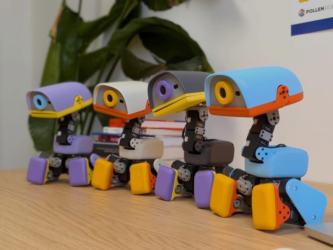 The image shows four robotic figures, each with a unique color scheme, arranged in a row on a light brown wooden desk. The first robot is purple and yellow, the second is white and orange, the third is gray and orange, and the fourth is blue and orange. Each robot has a camera-like head with a yellow beak and a small,