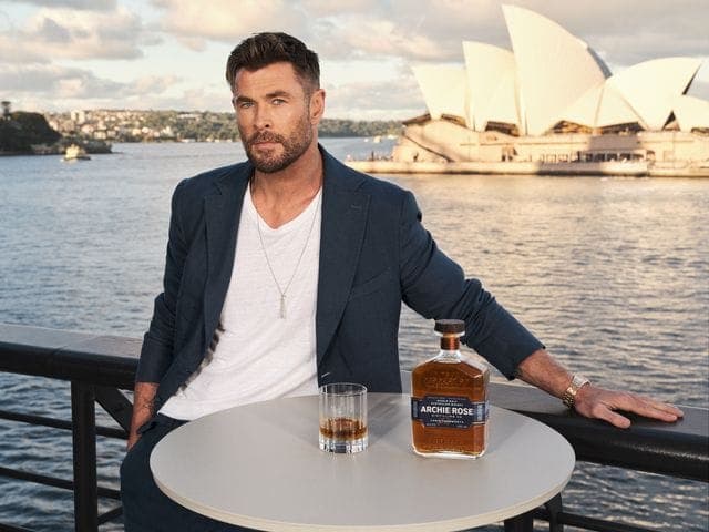 The image shows a man seated at a round, light gray table on a black railing overlooking a body of water. He is dressed in a navy blue blazer over a white t-shirt and a silver necklace. On the table, there is a glass of amber liquid, likely whiskey, and a bottle of Archie Rose whiskey. The iconic Sydney Opera House is