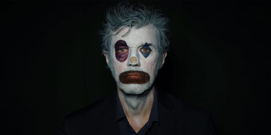The image shows a man with a painted face, possibly a clown, against a stark black background. His face is painted white with a large, dark purple circle on his left cheek, a small, light blue diamond on his right cheek, and a brown mustache. He is wearing a dark suit jacket and a dark-colored collared shirt. The man's