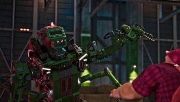 The image shows a large, green robot with a red head and arms, standing in a warehouse-like setting. The robot's arms are extended, and it appears to be holding a green object, possibly a tool or weapon. A person wearing a red and black plaid shirt is partially visible in the foreground, seemingly interacting with the