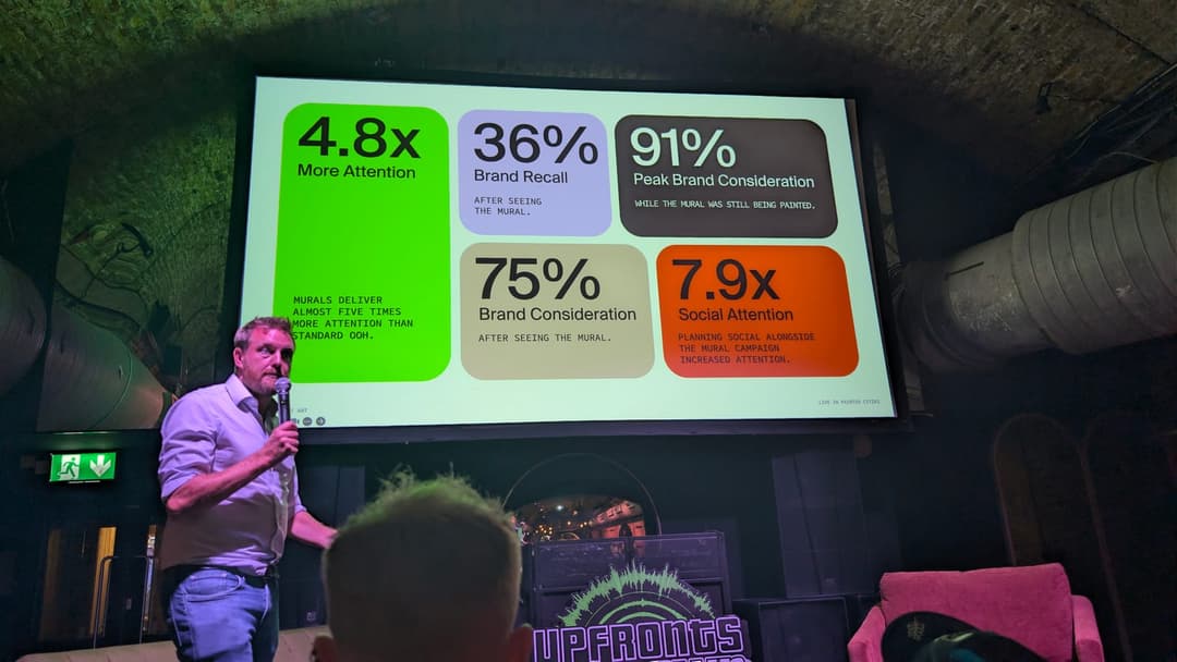 The image shows a man standing on a stage, holding a microphone and addressing an audience. He is wearing a light blue or grey shirt and blue jeans. Behind him, a large screen displays a graphic with a white background and black text. The graphic includes the following statistics: "4.8x More Attention", "36% Brand Reca