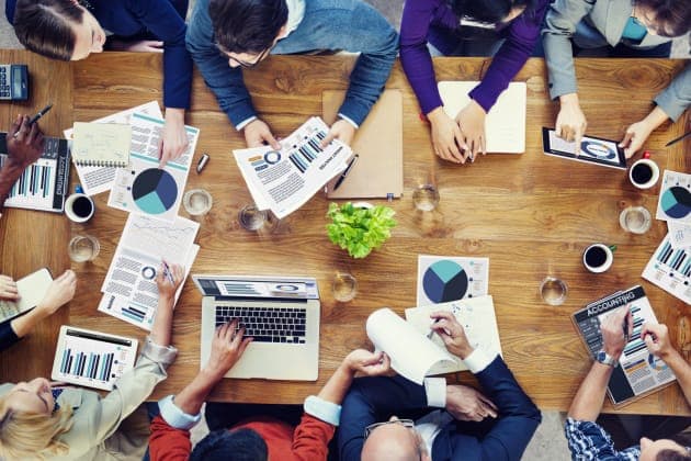 The image shows a group of people gathered around a wooden table, engaged in a meeting or brainstorming session. The table is covered with various papers, including charts and graphs, suggesting a discussion related to data analysis or financial planning. Some individuals are holding pens and notepads, while others are