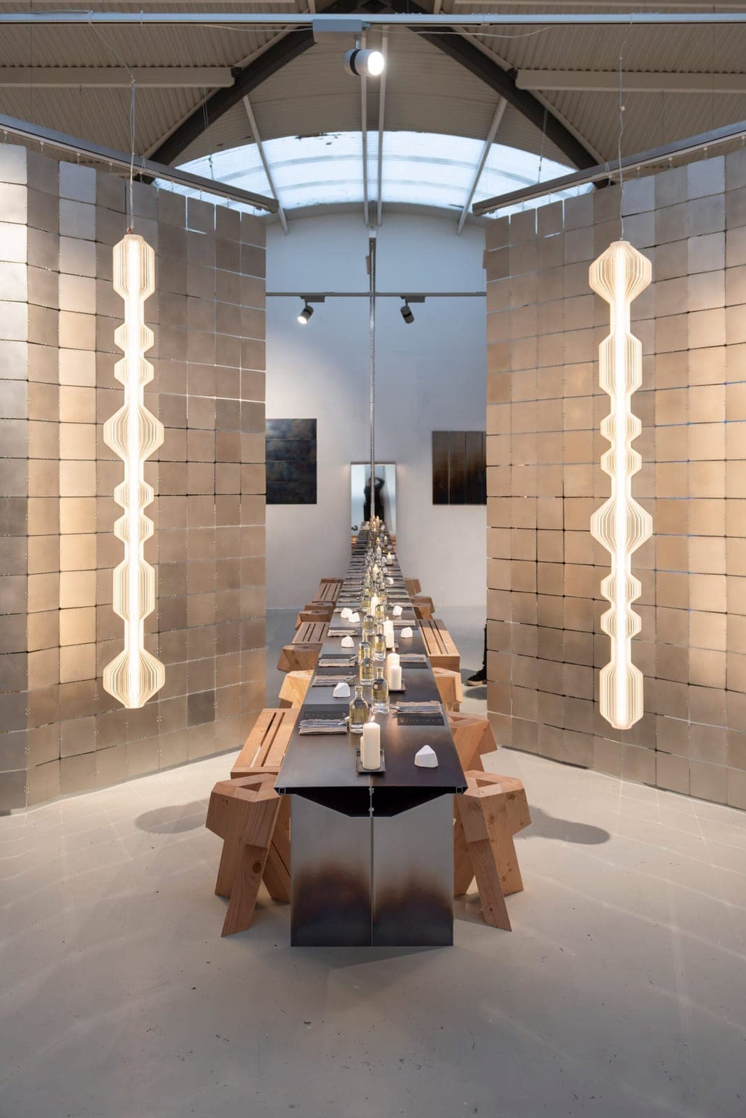 The image depicts a modern art gallery with a long, rectangular table as the centerpiece. The table is adorned with white candles and plates, and is surrounded by wooden stools. The walls are composed of large, rectangular, beige-colored blocks, creating a textured backdrop. Two tall, white, geometric pendant lights ha