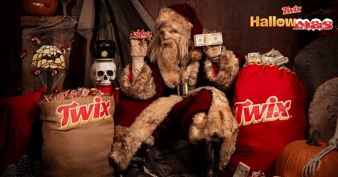 The image depicts a Halloween-themed scene featuring a figure dressed as Santa Claus. The figure is wearing a red suit with a fur-trimmed hood and a red hat, and is holding a Twix candy bar in each hand. The figure is seated on a dark brown or black surface, possibly a chair or couch. In the background, there are two b