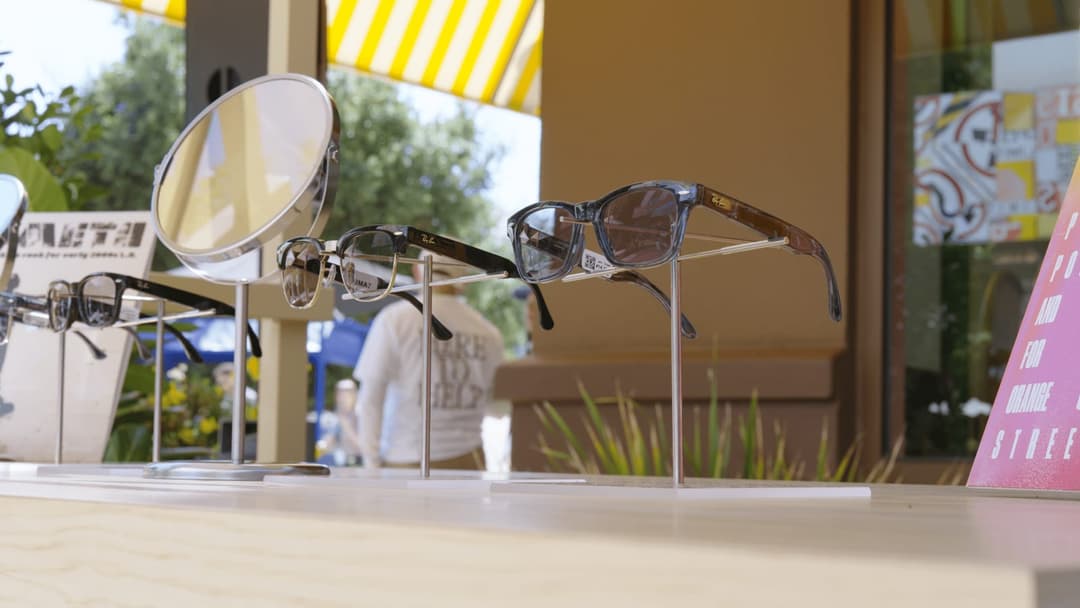 The image shows a display of sunglasses in a store window. The display consists of three pairs of sunglasses, each resting on a clear acrylic stand. The leftmost pair is black, the middle pair is dark brown or black, and the rightmost pair is dark brown or black. The sunglasses are arranged in a row, with the leftmost