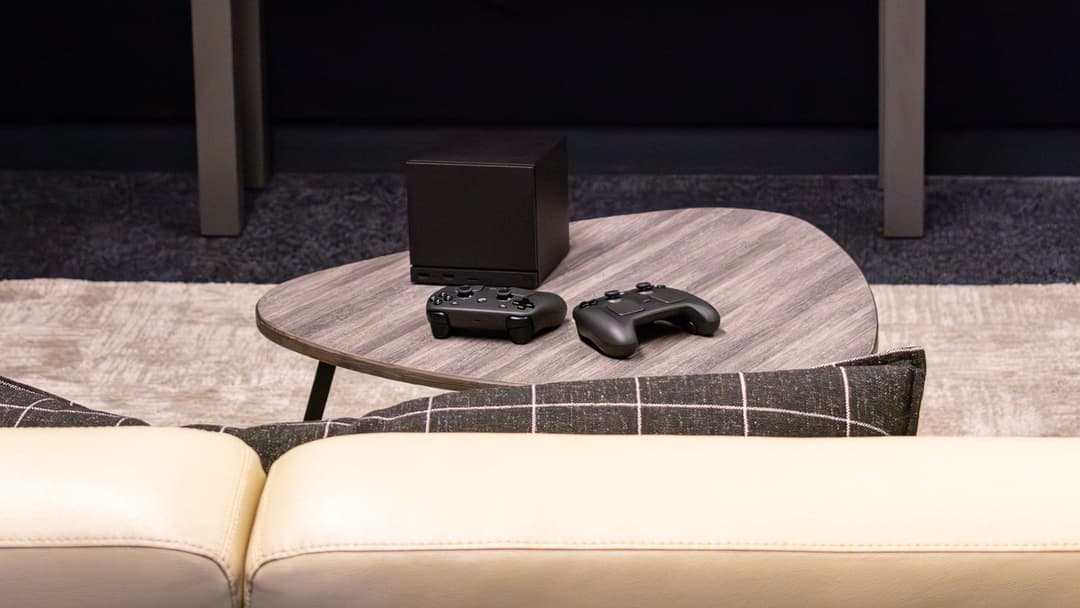 The image shows a cozy living room setting. A light brown wooden coffee table sits in the center of the room, with two black gaming controllers resting on it. A black speaker is positioned on the table, and a beige couch with a dark gray and white plaid throw pillow is visible in the foreground. The room features a dar