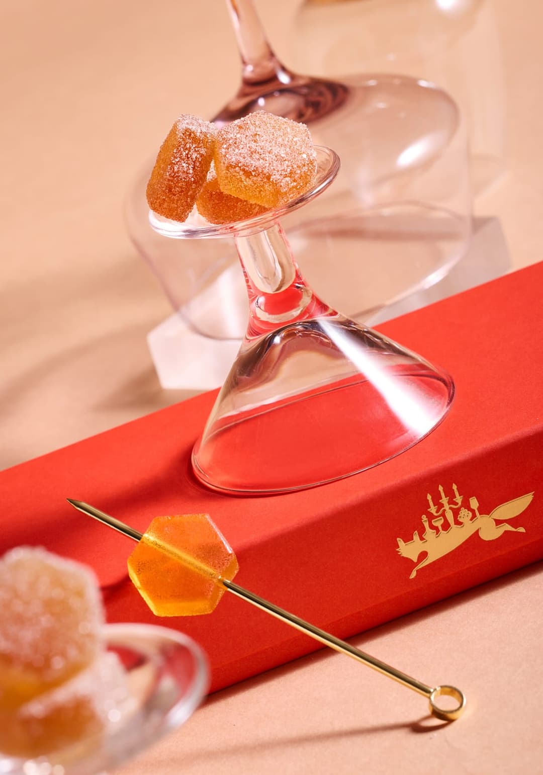 The image shows a collection of objects arranged on a beige surface. A clear glass cocktail glass with a pinkish-purple stem is positioned in the center, holding a small pile of orange-colored candies dusted with white sugar. To the right of the glass, a gold cocktail stirrer with a small, gold-colored hole at the end