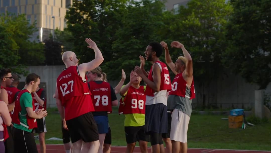 The image captures a group of athletes, possibly a track team, celebrating on a track. The athletes are wearing red jerseys with white numbers, and some are wearing black shorts. The track is surrounded by a grassy area, and a building is visible in the background. The athletes are raising their arms in the air, sugges