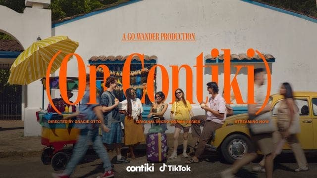 A group of people walks along a sunlit street past a white building with a yellow-and-white striped umbrella, with overlaid orange text reading "(On Contiki)", "A GO WANDER PRODUCTION", and "Directed by Gracie Otto", plus the Contiki and TikTok logos.