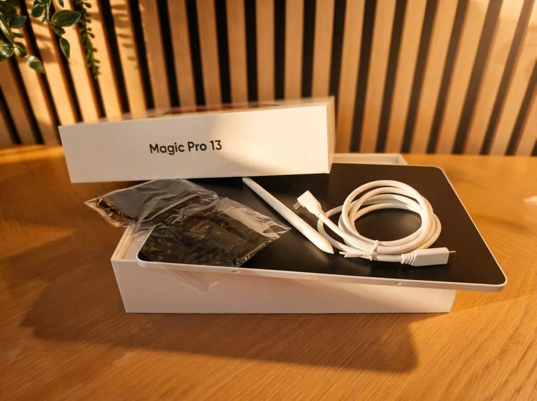 The image shows a white box labeled "Magic Pro 13" resting on a wooden surface. The box contains a black tablet, a white stylus, a white cable, and a black plastic bag. The stylus is positioned on top of the box, and the cable is connected to the stylus. The tablet and stylus are placed on a black surface, which appear