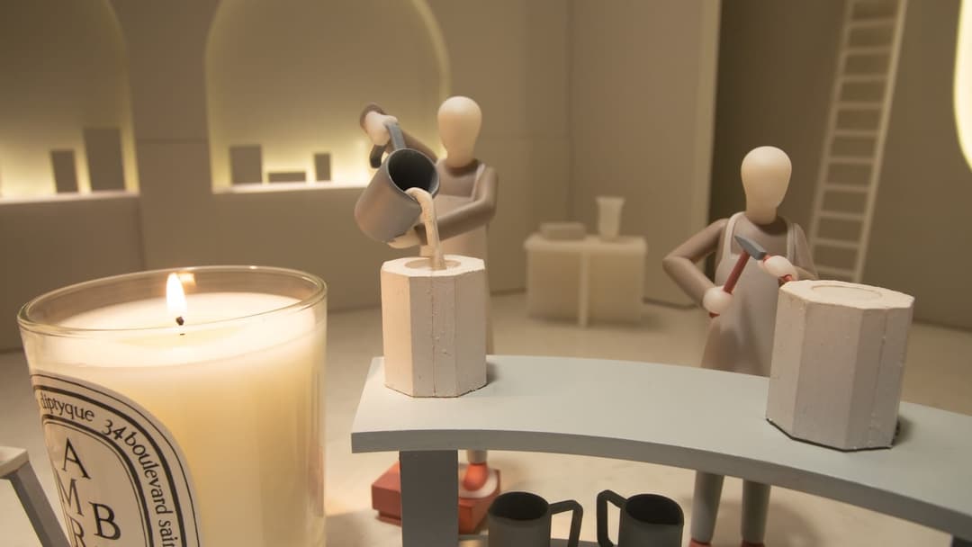 The image depicts a miniature scene set in a room with a white ceiling and walls. The room features a curved table with a gray surface, where three small, white, humanoid figures are engaged in a crafting activity. One figure is pouring a liquid from a gray pitcher into a hexagonal mold, while another holds a red tool.