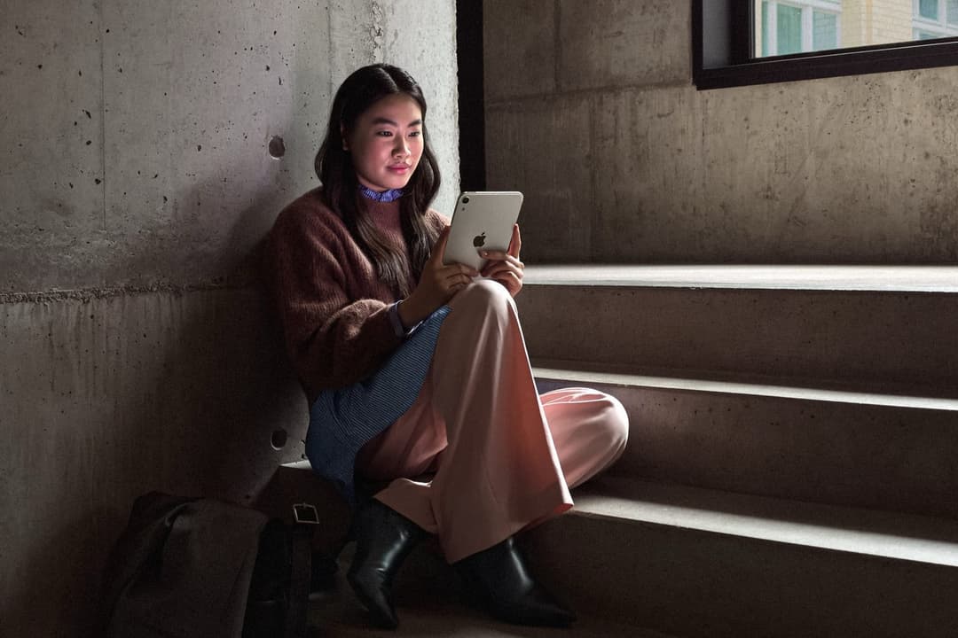A young woman with long, dark hair is seated on a concrete staircase, engrossed in her white Apple iPad. She is dressed in a brown sweater, a blue and white striped shirt, and pink pants, and is wearing black boots. A black backpack rests beside her on the stairs. The staircase is set against a concrete wall, and a win