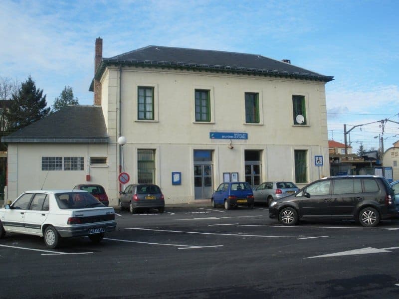 The image shows a two-story building with a cream or off-white facade and a dark gray roof. The building has a sign on the front that reads "Bruyiers-Légales" and features several windows with dark green or black frames. The building is situated in a parking lot with several cars parked in front, including a white car,