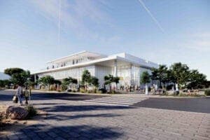 Daytime exterior of a modern white building with a glass facade and tall windows, with trees and a paved plaza in front. The sky is blue with faint contrails. No legible text is visible.