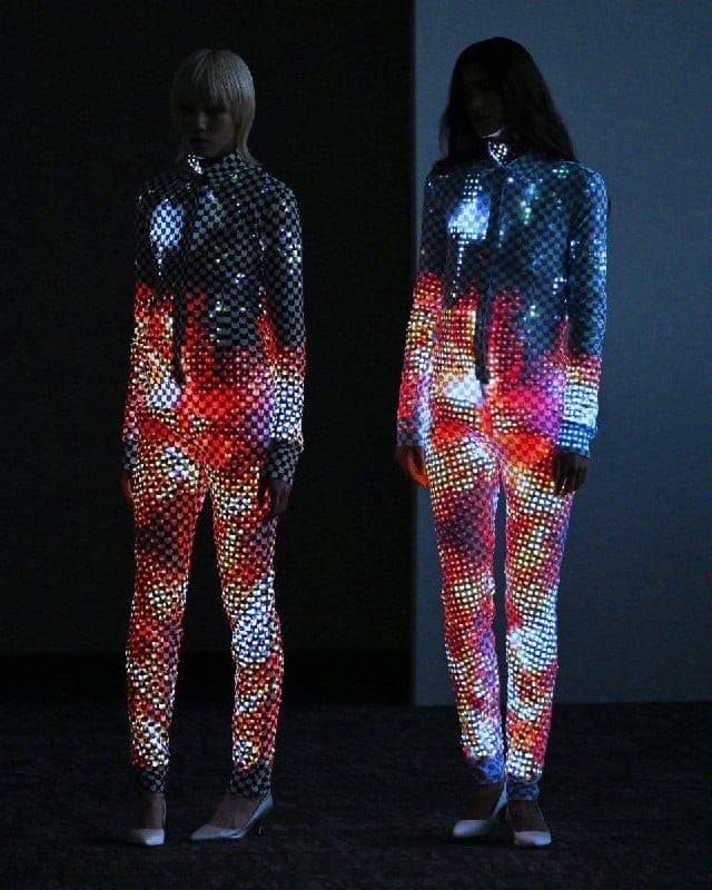 The image shows two individuals standing side by side in a dimly lit room. The person on the left is wearing a black and red checkered jumpsuit with a zipper down the front, and the person on the right is wearing a blue and pink checkered jumpsuit with a zipper down the front. Both individuals are wearing white shoes.