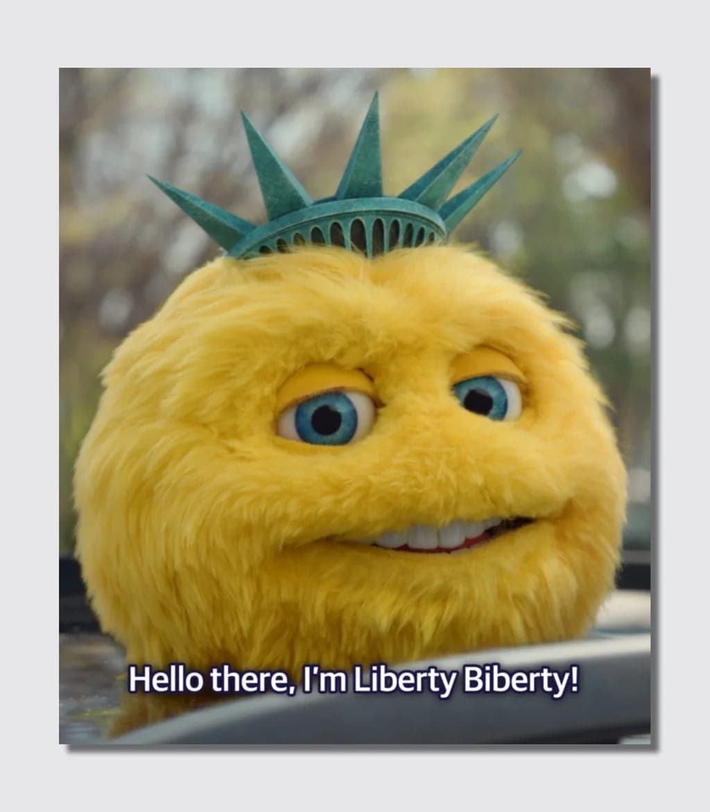 A still of a fuzzy yellow creature wearing a green Statue of Liberty crown peeks over a car hood, with the subtitle 'Hello there, I'm Liberty Biberty!'.
