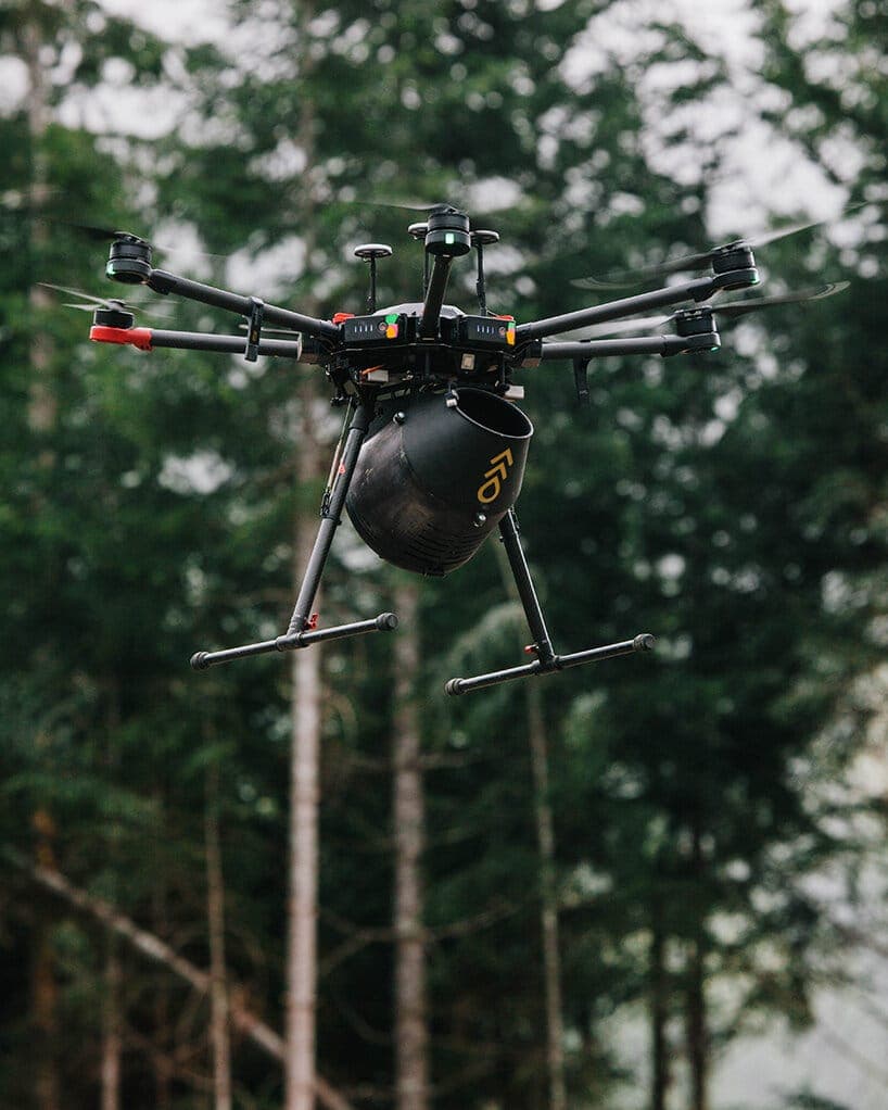 The image shows a black drone with six propellers, equipped with a camera and a black bucket attached to its underside. The drone is flying in a forested area, with tall trees forming a natural canopy in the background. The drone's body is primarily black with some red and yellow accents. The drone's propellers are blu