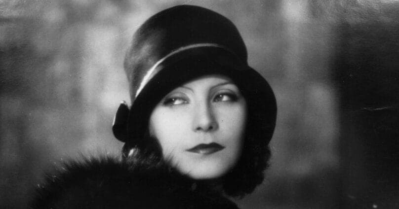 This black and white photograph captures a woman with a serious expression, wearing a stylish hat and a fur-trimmed coat. The woman's gaze is directed off to the side, and her lips are slightly parted. The background is out of focus, creating a sense of depth and drawing attention to the woman. The photograph is in bla