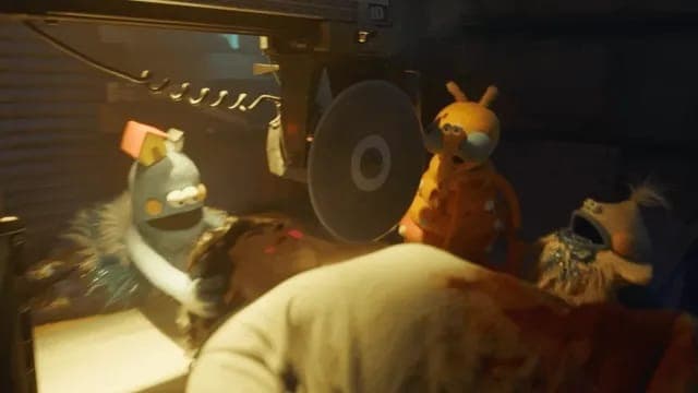 The image shows a scene that appears to be from a movie or animation, set in a dimly lit room. A large, round, dark-colored object, possibly a speaker or a piece of equipment, dominates the center of the image. To the left, a blue, furry creature with a red/orange object on its head is interacting with a dark, possibly