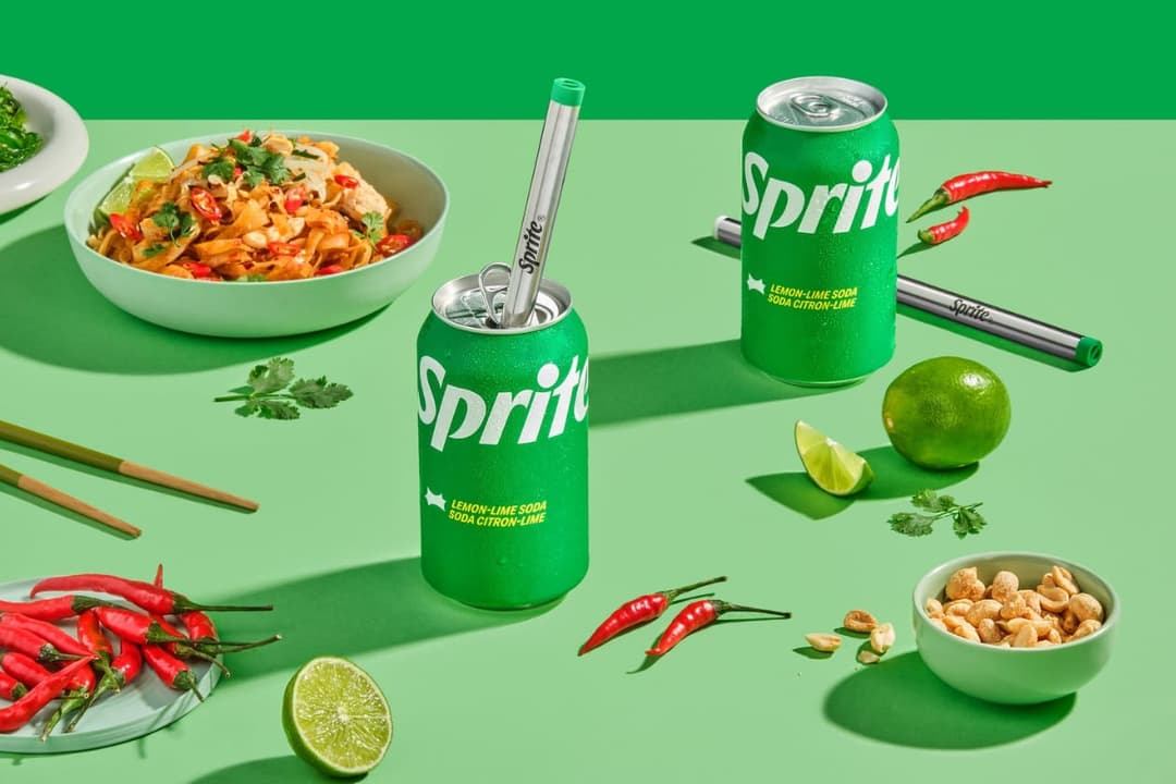 The image presents a vibrant still life scene set against a solid green background. At the center is a green Sprite can, featuring a silver straw and a lime green label with the word "Sprite" prominently displayed. To the left of the Sprite can is a white bowl filled with noodles, garnished with green herbs and topped