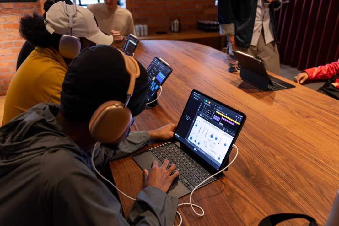 The image shows a group of people gathered around a wooden table, engaged in a collaborative activity. The table is adorned with several laptops, including a large Apple MacBook, a smaller tablet, and a smaller laptop. The laptops are open and displaying various screens, suggesting a collaborative project or discussion