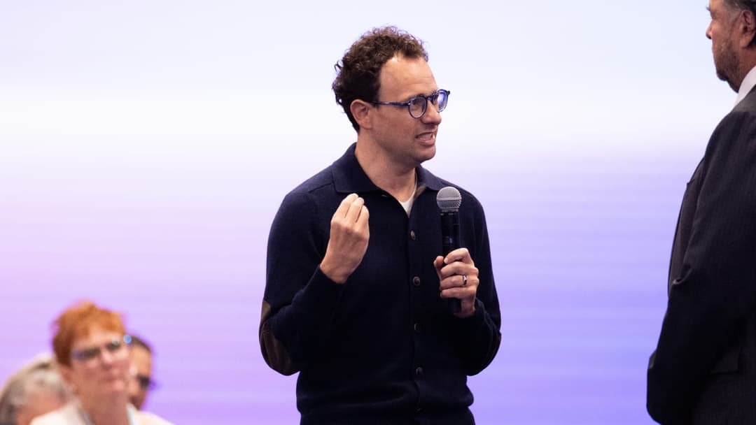 The image shows a man standing on a stage, holding a microphone and gesturing with his right hand. He is wearing a dark blue cardigan over a white shirt and glasses. The stage has a purple background. In the background, a portion of a seated audience is visible, and a man in a dark suit is standing to the right of the