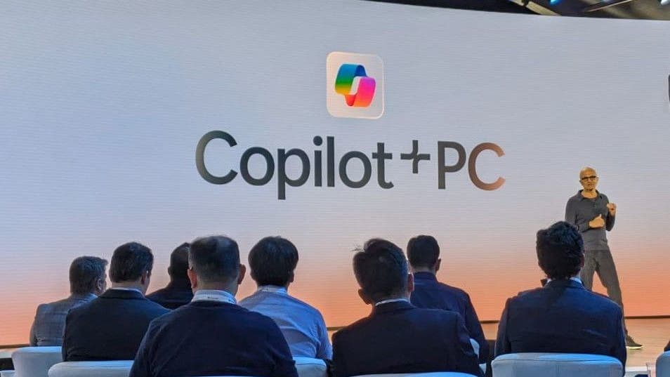 The image captures a presentation taking place on a stage. A man in a gray shirt and dark pants is standing on the stage, gesturing with his hands as he speaks. The stage is set against a white backdrop with a gradient of orange and pink tones. In the foreground, a group of people are seated in rows of chairs, attentiv